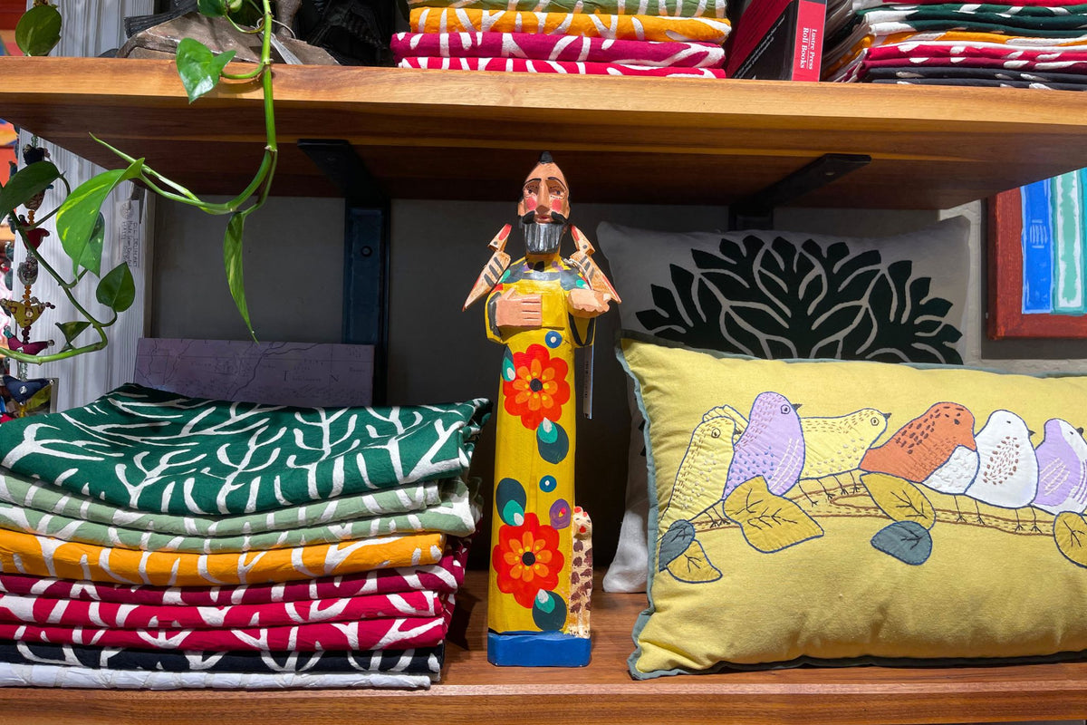 A colorful hand-painted wooden figurine of Saint Francis of Assissi stands on a wooden shelf at Hill Station in Ashland, between a stack of folded Tree of Life Wall Hangings and a lime green pillow with birds on a branch appliqued onto it.