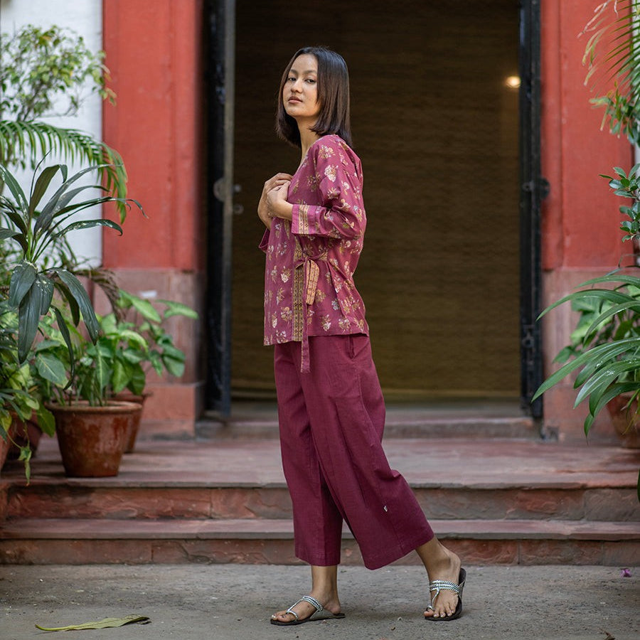Woman in a plum colored top and maroon pants standing in front of a brick red building entrance.
