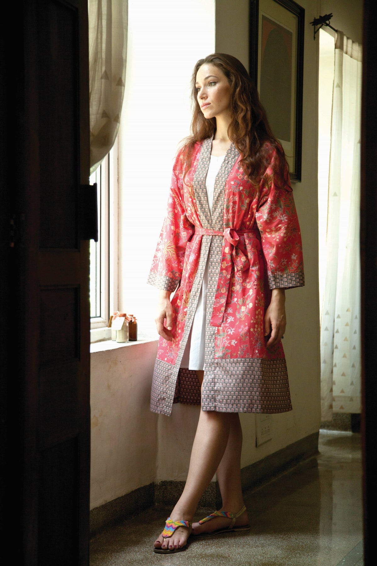 A woman wearing a pink and red patterned robe over a white dress, standing in a doorway in a sunlit room.