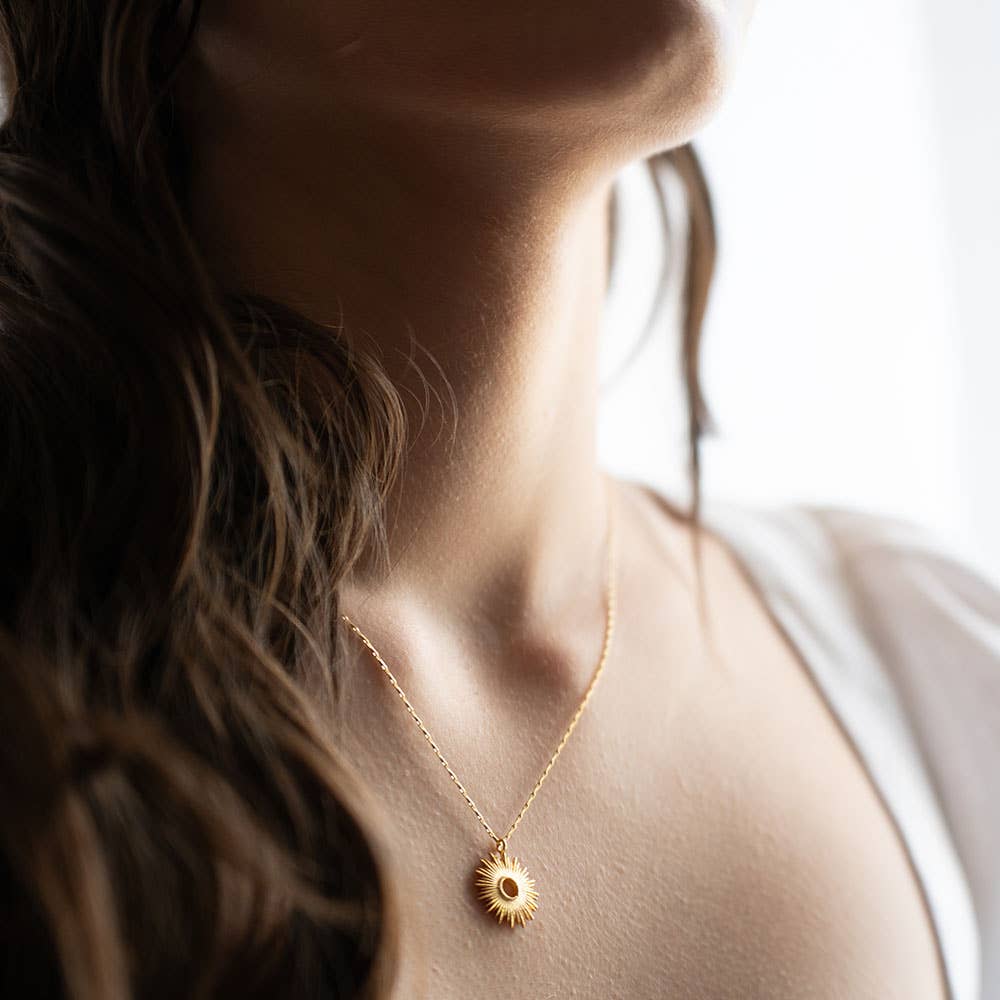 Gold sunburst pendant necklace on delicate chain worn by woman wearing an off-white top. Shadowed light.